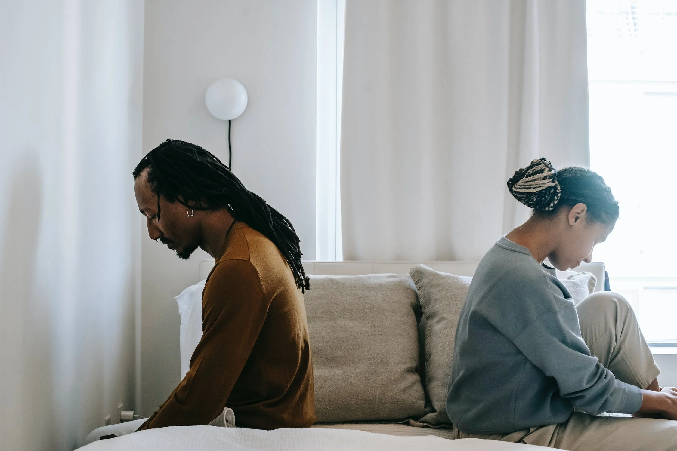 A troubled African American couple sits on a bed, their expressions reflecting confusion and misunderstanding, as they grapple with an emotional moment in their relationship.